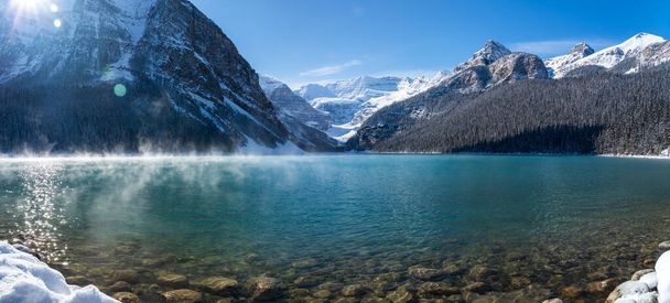 Λίμνη Louise νωρίς το χειμώνα ηλιόλουστη μέρα το πρωί. Mist επιπλέουν σε τυρκουάζ επιφάνεια του νερού χρώμα. Καθαρός γαλάζιος ουρανός, χιονισμένα βουνά στο βάθος. Όμορφο φυσικό τοπίο στο Banff National Park. - Φωτογραφία, εικόνα