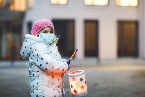 聖マーティンの行列のための自作ランタンを保持幼児の女の子.子供は医療マスクを着用する。幼稚園の子供や家族のパレードです。ドイツの伝統マルティンスムツーク流行コロナウイルスの隔離 - 写真・画像