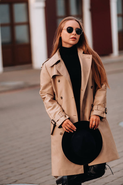 Stylish young woman in a beige coat and a black hat in his hands and glasses on a city street. Women's street fashion. Autumn clothing.Urban style - Foto, afbeelding