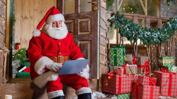 Santa Claus is sitting on the porch of the house and uses a laptop. Christmas concept - Foto, afbeelding