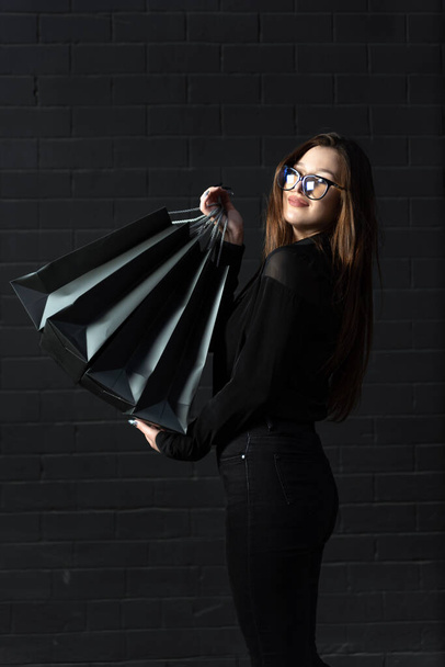 Молодая девушка в черной одежде держит сумки для покупок у черной стены. Вертикальная рамка. - Фото, изображение