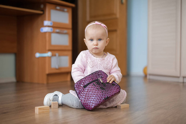 A one-year-old baby in a cute pink dress with a tulle skirt sits on the floor with a purple bag. - Foto, afbeelding
