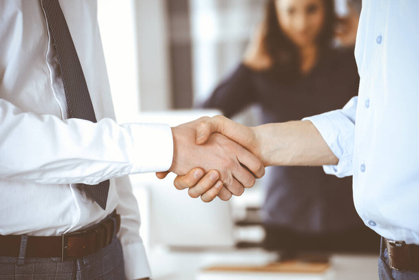 Two businessmen are shaking hands in office, close-up. Happy and excited business woman stands with raising hands at the background. Business people concept - Photo, Image