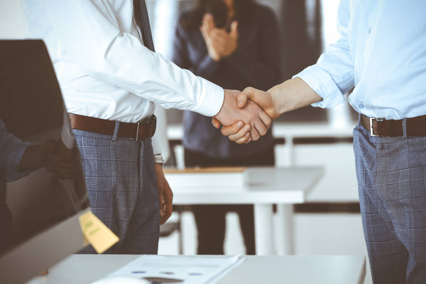 Two businessmen are shaking hands in office, close-up. Happy and excited business woman stands with raising hands at the background. Business people concept - 写真・画像