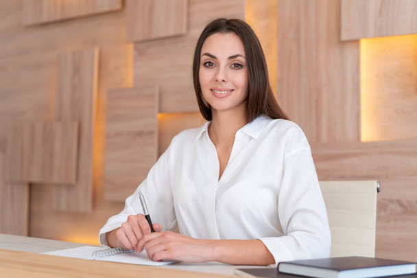 Office manager woman in white shirt, beautiful smile, sitting to table with notebook in wooden office interior, side view. Friendly office secretary looking at the camera. Concept of work - Valokuva, kuva