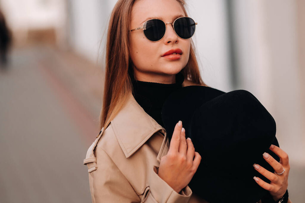 Stylish young woman in a beige coat and a black hat in his hands and glasses on a city street. Women's street fashion. Autumn clothing.Urban style - Φωτογραφία, εικόνα