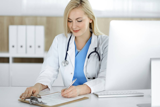Woman-doctor at work while sitting at the desk in hospital or clinic. Blonde cheerful physician filling up medication history record form - Фото, изображение