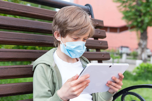 A boy with medical blue mask sitting on the street bench with tablet computer - Foto, immagini