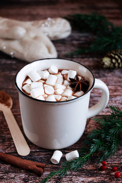 White cup of hot spiced cacao or chocolate with marshmallows on a rustic wooden tabletop with fir branches and knitted gloves in the background.  - Foto, imagen