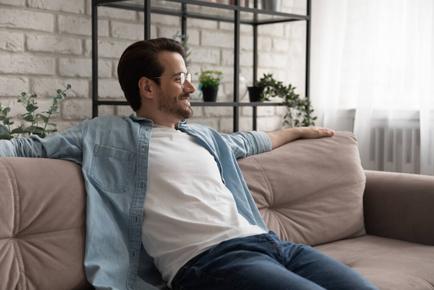 Smiling young handsome man in eyeglasses relaxing on comfortable couch. - 写真・画像