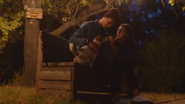 Romantisch jong paar met jack russell terrier hond zit op bank in het nachtpark - Video