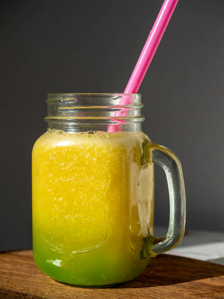 vaso di vetro illuminato dal sole con frullati gialli. C'e 'un tubo per bere nel barattolo. Sfondo in legno - Foto, immagini