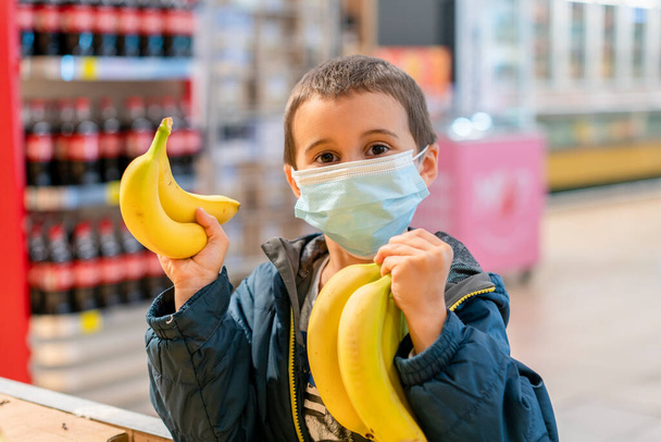 Παιδί που φοράει χειρουργική μάσκα προσώπου αγοράζοντας φρούτα σε σούπερ μάρκετ σε πανδημία κορωναϊού. Το αγοράκι στο σούπερ μάρκετ φοράει ιατρική μάσκα. Καραντίνα από τον ιό του στέμματος. τονισμένο. - Φωτογραφία, εικόνα