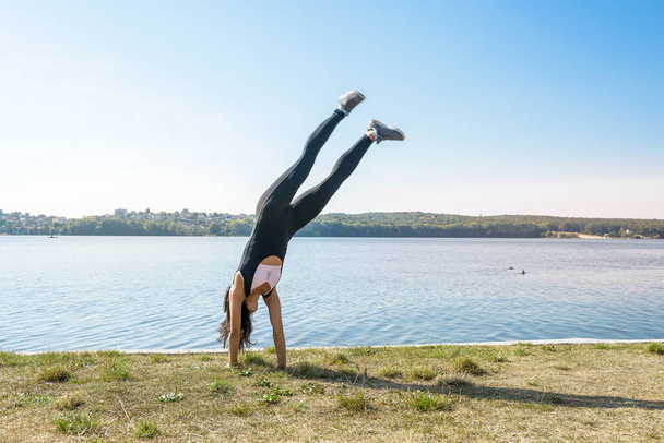 笑顔の若い女性の体操選手は、湖の近くの屋外で分割にジャンプしています。健康的なライフスタイル. - 写真・画像