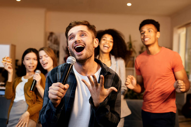 Leuk thuis. Enthousiaste knappe jonge man zingen met microfoon tijdens het spelen van karaoke met vrienden binnen, staan in het moderne appartement - Foto, afbeelding