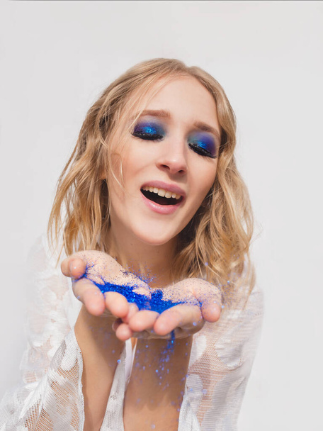 Pretty young cheerful blonde blowing blue glitter from her hands. Make up, fashion, magic, holidays, miracle concept - Фото, изображение