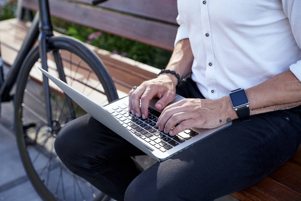 Trabajadora ocupada. Foto recortada de hombre de negocios moderno de mediana edad en camisa blanca trabajando en su computadora portátil mientras está sentado en el banco al aire libre con una bicicleta a su lado - Foto, Imagen