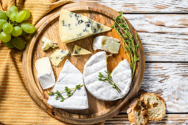 Assorted cheeses on a round wooden cutting Board. Camembert, brie and blue cheese with grapes. White wooden background. Top view. - Foto, Imagen