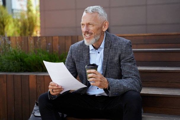 Trouve de nouveaux moyens. Élégant homme d'affaires d'âge moyen tenant des papiers, prenant une pause tout en étant assis avec une tasse de café à l'extérieur - Photo, image