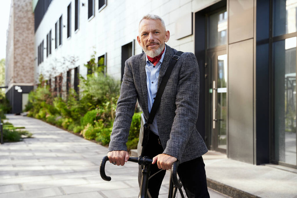 Zelfverzekerde volwassen zakenman kijkend naar camera terwijl hij overdag met zijn fiets buiten staat - Foto, afbeelding