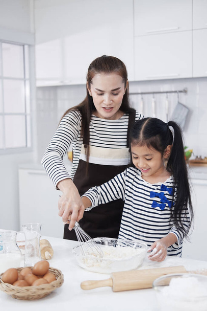 笑顔アジアの母親と小さなアジアの女の子の子供は台所で一緒に木製のテーブルの上にパン屋を焼くために料理しています.自家製のペストリーとパンのための混合成分。家族の愛と家庭学校のコンセプト. - 写真・画像