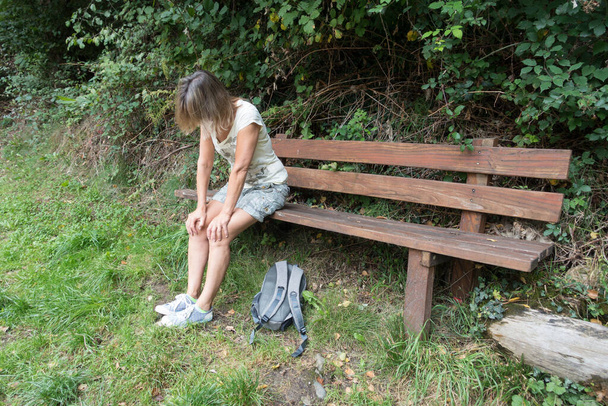 Naiskävelijä istuu puisella penkillä kädet polvillaan väsyneenä. Reppu makaa ruohikolla hänen jalkojensa juuressa.. - Valokuva, kuva