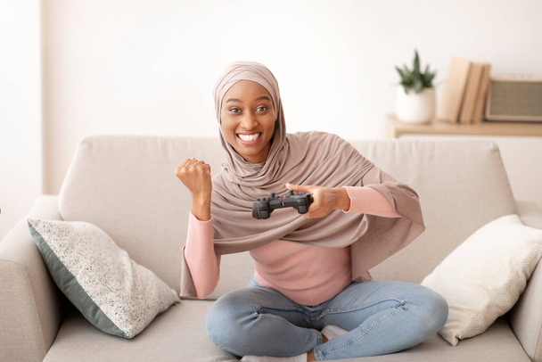 Wochenendvergnügen. Glückliche schwarze Frau im Hijab mit Steuerknüppel spielt drinnen Videospiele und macht JA-Geste - Foto, Bild
