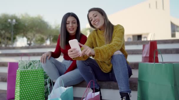 Két fiatal gyönyörű nő ül a téren lépéseket beszél vásárlás online színes táskák - Egy pár mosolygós női fogyasztók szórakozás szabadtéri okostelefon használatával - Fókusz a telefonra - Felvétel, videó