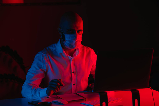Łysy facet w masce medycznej pracuje zdalnie na laptopie w domu. Facet gapi się podczas notatek. Mężczyzna z długopisem przed komputerem pod niebieskim i czerwonym światłem. - Zdjęcie, obraz