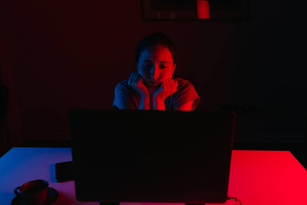 Een vrouw werkt thuis op afstand op een laptop. Een meisje rust haar hoofd op haar handen die op de tafel leunt. Een vrouwelijke werknemer verveelt zich voor de computer onder blauw en rood licht. - Foto, afbeelding