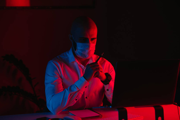 Ένας φαλακρός με μάσκα ιατρικού προσώπου δουλεύει εξ αποστάσεως σε ένα λάπτοπ στο σπίτι. Ένας τύπος διαβάζει και κάνει σημειώσεις. Ένας άντρας υπάλληλος με τα χέρια του διπλωμένα μπροστά στον υπολογιστή κάτω από μπλε και κόκκινο φως.. - Φωτογραφία, εικόνα