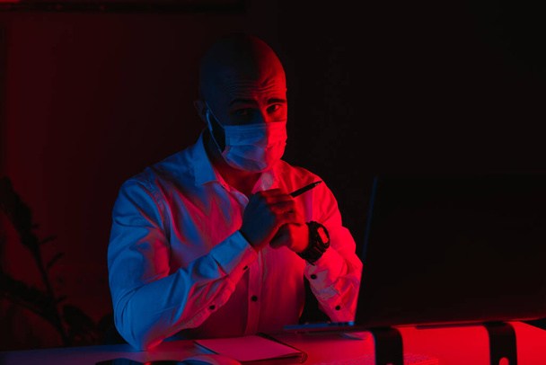 Ένας φαλακρός με μάσκα ιατρικού προσώπου δουλεύει εξ αποστάσεως σε ένα λάπτοπ στο σπίτι. Ένας τύπος κοιτάζει επίμονα. Ένας άντρας υπάλληλος με στυλό μπροστά στον υπολογιστή κάτω από μπλε και κόκκινο φως.. - Φωτογραφία, εικόνα