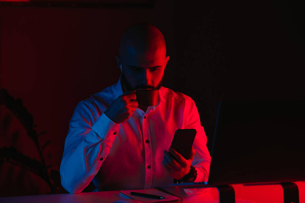 Un uomo con la barba lavora a distanza su un portatile a casa. Un tizio con le cuffie beve caffe 'e legge i notiziari. Un dipendente di fronte al computer nei raggi di luce blu e rossa. - Foto, immagini