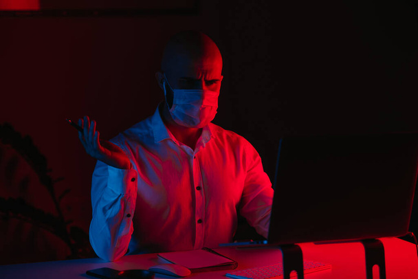 Ein glatzköpfiger Mann mit medizinischer Gesichtsmaske arbeitet zu Hause an einem Laptop. Ein Mann wird von Missgunst überrascht. Ein männlicher Mitarbeiter mit einem Stift vor dem Computer unter blauem und rotem Licht. - Foto, Bild