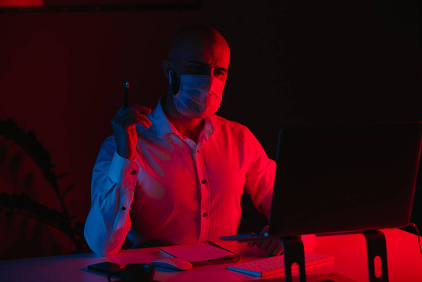 Egy kopasz férfi orvosi maszkban távirányítással dolgozik egy laptopon otthon. Egy fickó olvas és jegyzetel. Egy férfi alkalmazott a számítógép előtt, kék és piros fény alatt.. - Fotó, kép