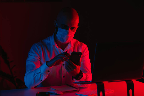 Ein glatzköpfiger Mann mit medizinischer Gesichtsmaske arbeitet zu Hause an einem Laptop. Ein Mann liest die Nachrichten auf einem Smartphone. Ein männlicher Mitarbeiter mit einem Stift vor dem Computer unter blauem und rotem Licht. - Foto, Bild