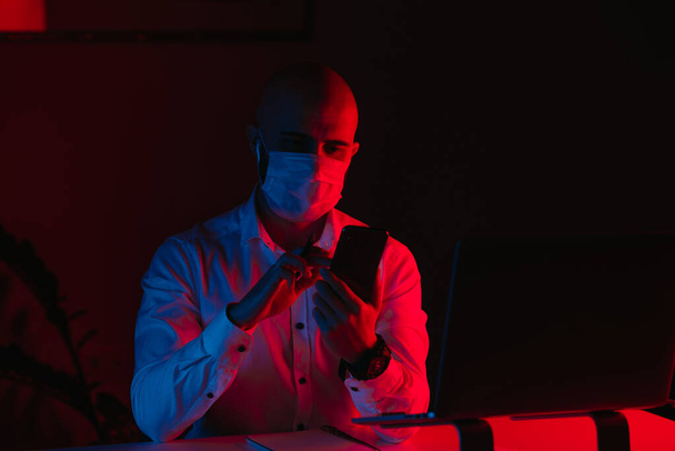 Un hombre calvo con una mascarilla médica está trabajando remotamente en un portátil en casa. Un tipo con auriculares está leyendo las noticias en un teléfono inteligente. Un empleado masculino frente a la computadora bajo la luz azul y roja. - Foto, Imagen