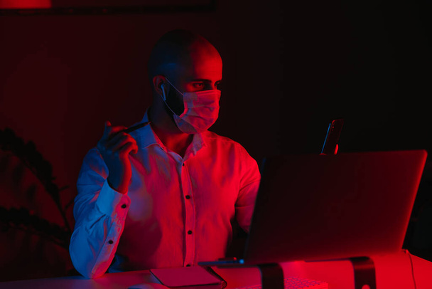 Ένας φαλακρός με μάσκα ιατρικού προσώπου δουλεύει εξ αποστάσεως σε ένα λάπτοπ στο σπίτι. Ένας τύπος με ακουστικά κρατάει ένα smartphone. Ένας άντρας υπάλληλος με στυλό μπροστά στον υπολογιστή κάτω από μπλε και κόκκινο φως.. - Φωτογραφία, εικόνα