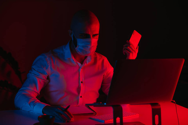 Ein glatzköpfiger Mann mit medizinischer Gesichtsmaske arbeitet zu Hause an einem Laptop. Ein Mann mit Kopfhörern hält ein Handy in der Hand. Ein männlicher Mitarbeiter mit einem Stift vor dem Computer unter blauem und rotem Licht. - Foto, Bild