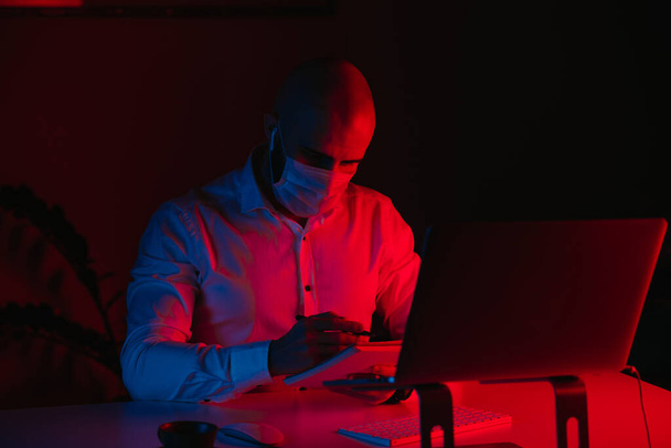 Un chauve dans un masque médical travaille à distance sur un ordinateur portable à la maison. Un type avec des écouteurs prend des notes. Un employé masculin avec un stylo devant l'ordinateur sous la lumière bleue et rouge. - Photo, image