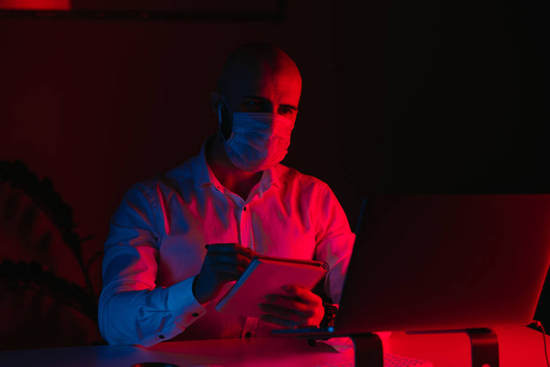 Ένας φαλακρός με μάσκα ιατρικού προσώπου δουλεύει εξ αποστάσεως σε ένα λάπτοπ στο σπίτι. Ένας τύπος με ακουστικά ελέγχει τις σημειώσεις του. Ένας άντρας υπάλληλος με στυλό μπροστά στον υπολογιστή κάτω από μπλε και κόκκινο φως.. - Φωτογραφία, εικόνα