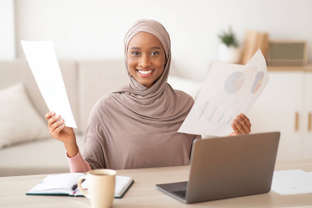 Positive schwarze Muslimin im Hidschab arbeitet von zu Hause aus und hält Dokumente in der Nähe von Laptop-Computern - Foto, Bild