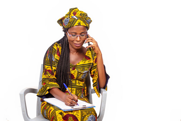 Взрослая деловая женщина сидит улыбаясь и разговаривая по телефону во время записи в блокноте - Фото, изображение