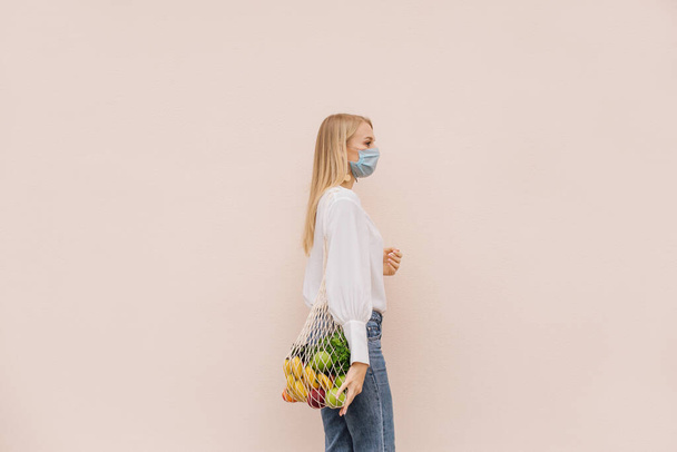 Young woman wearing protective face mask for prevention from coronavirus Covid-19 pandemic holding string shopping bag with fruits. Eco lifestyle. Conscious consumption. New normal. Copy space - Fotografie, Obrázek