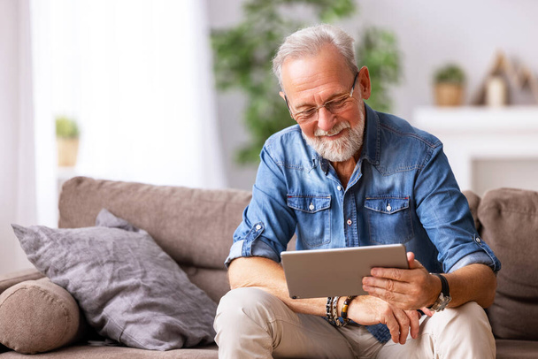 Счастливый старший бородатый мужчина в повседневной одежде просматривает планшет и улыбается, сидя на диване в доме - Фото, изображение