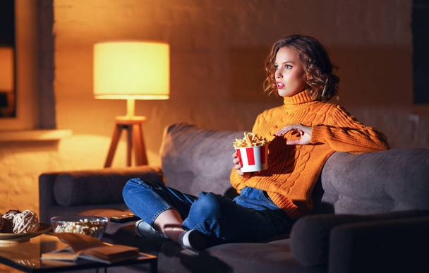 Nuori mykistynyt nainen syö ranskalaisia perunoita ja katselee jännittävää toimintapakkaista elokuvaa televisiosta kotona illalla alon - Valokuva, kuva