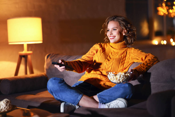 young  cheerful woman eating popcorn and watches  movie on  cable TV while switching channels with the remote control at home in evening  alon - Zdjęcie, obraz