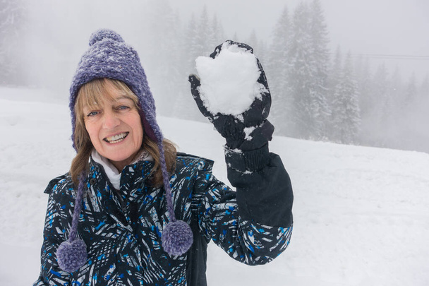 A lady holds a snowball in a gloved hand.She is laughing and looks to be about to throw it towards camera.She wears a purple woollen hat with tassels and baubles and a ski jacket.Snow and trees in a misty winter background. - Photo, Image