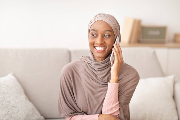 Sonriente dama negra en hijab hablando en el teléfono inteligente en casa - Foto, imagen