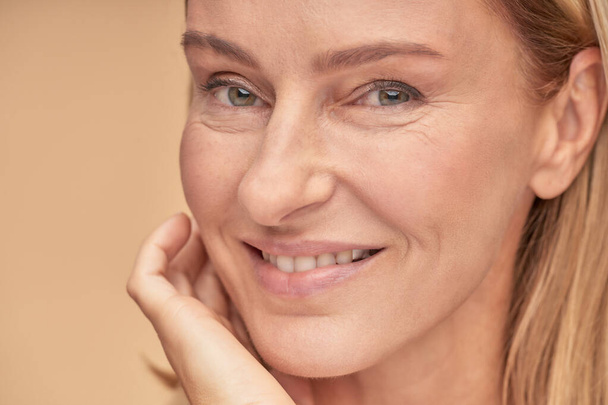 Anti aging péče o pleť. Detail portrét krásné ženy středního věku dotýká její tvář a usmívá se na kameru, zatímco pózuje přes béžové pozadí ve studiu - Fotografie, Obrázek